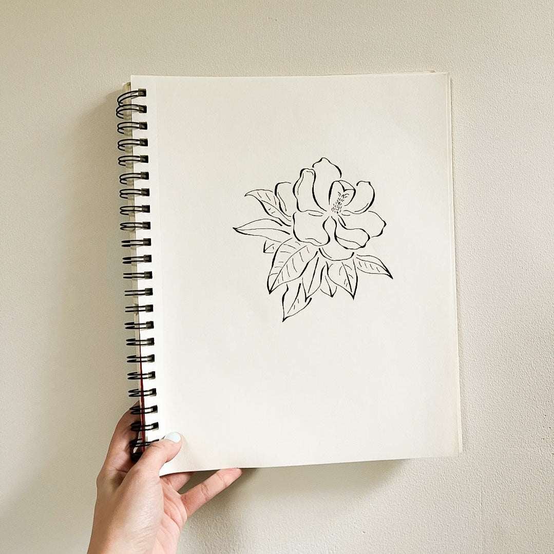 A hand holding a white spiral-bound sketchbook with a simple black line drawing of a flower on the cover, set against a plain beige wall.