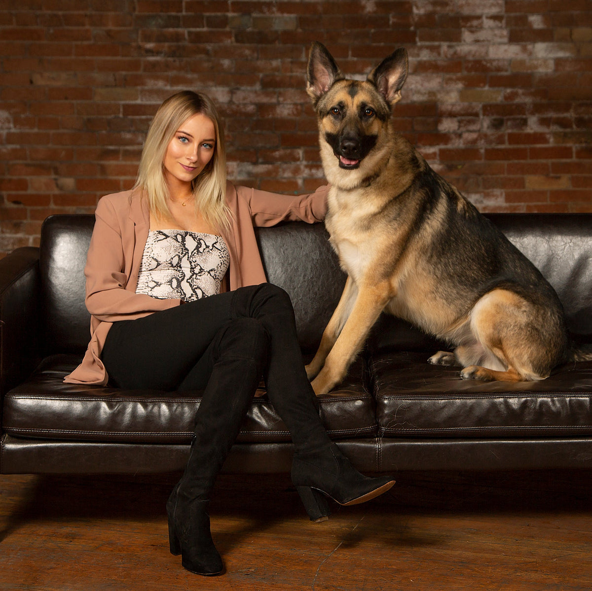 A woman sitting on a black leather couch next to a German Shepherd dog, posed in front of a rustic brick wall background.
