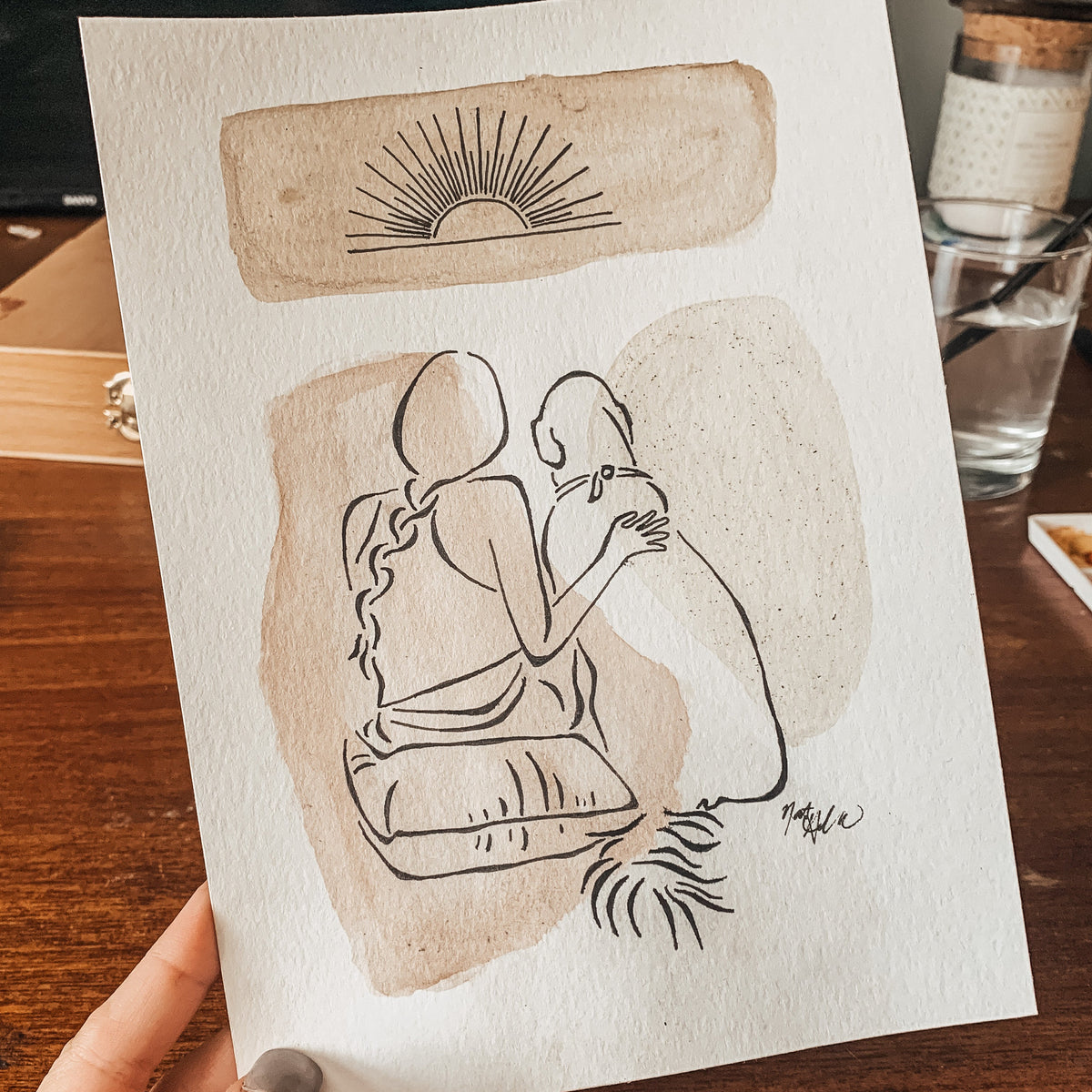 A hand holds a square art print featuring a minimalist black line drawing of a seated couple on beige watercolor washes, with a sun motif at the top, against a warm wooden tabletop background.