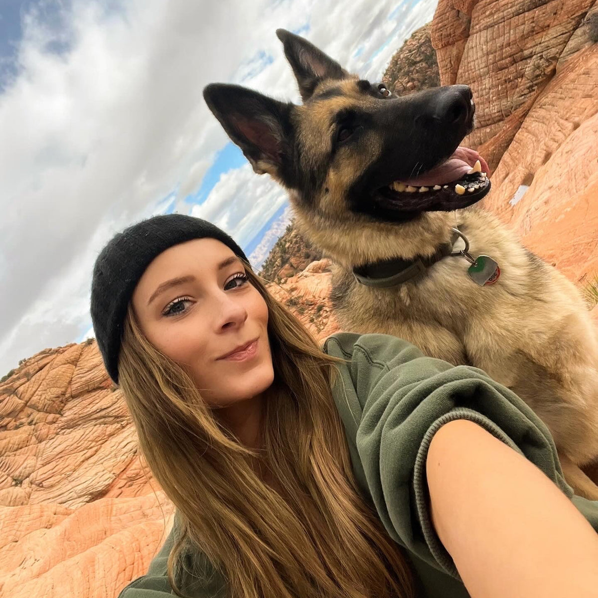 A woman taking a selfie with a German Shepherd dog in a desert canyon setting. The woman is wearing a black beanie and an olive green sweatshirt, while the dog has a black collar with a green tag.