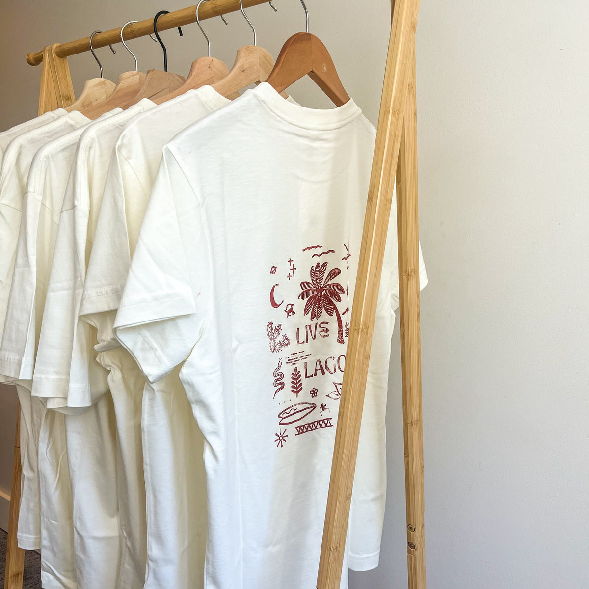 A white short-sleeve graphic T-shirt hanging on a wooden clothing rack, with other white shirts behind it. The shirt features a reddish-brown tropical print with palm trees and the words 'LIVE LAGOM'.