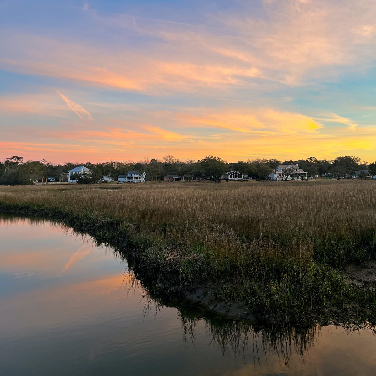 A scenic landscape photograph of a marshland at sunset, featuring a calm body of water in the foreground reflecting the sky, golden reeds, and a line of houses in the distance under a blue, orange, and yellow sunset sky.
