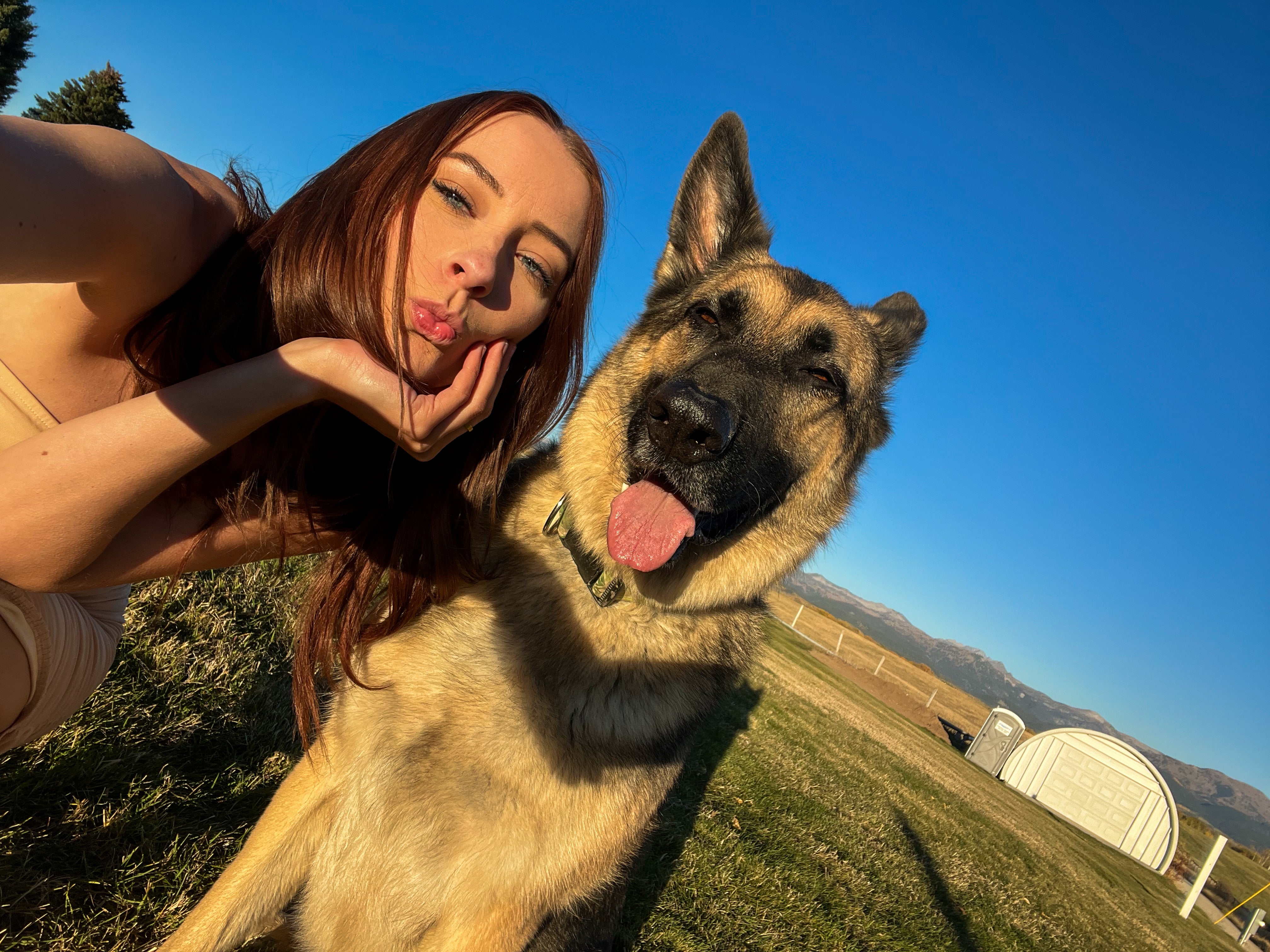 A woman posing outdoors with a German Shepherd dog under a bright blue sky. The woman is on the left, making a face, and the dog is on the right, looking at the camera with its tongue out.