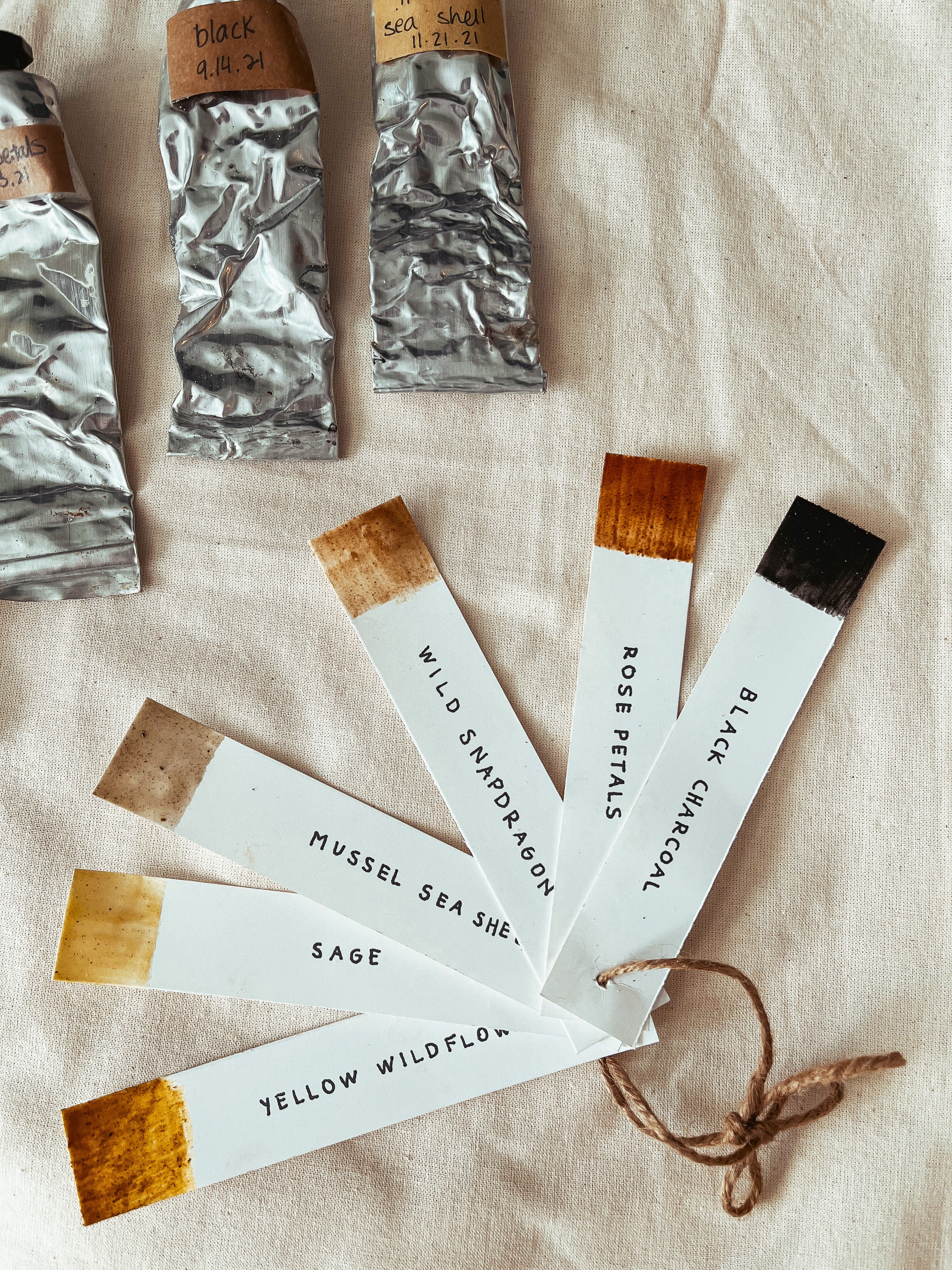 A styled flat-lay of watercolor paint tubes and matching color chips on a beige linen background. The tubes are wrapped in silver foil with handwritten labels, and several white color swatch cards with painted wood-tone tips are fanned out, showing colors like Yellow Wildflower, Mossel Sea Sheaf, Black Charcoal, Rose Petals, and Wild Syntheton.