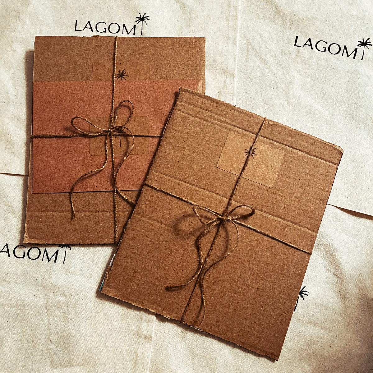 Two brown cardboard mailers wrapped in twine with a small black palm tree graphic, placed on top of white canvas bags printed with the brand name LAGOM.