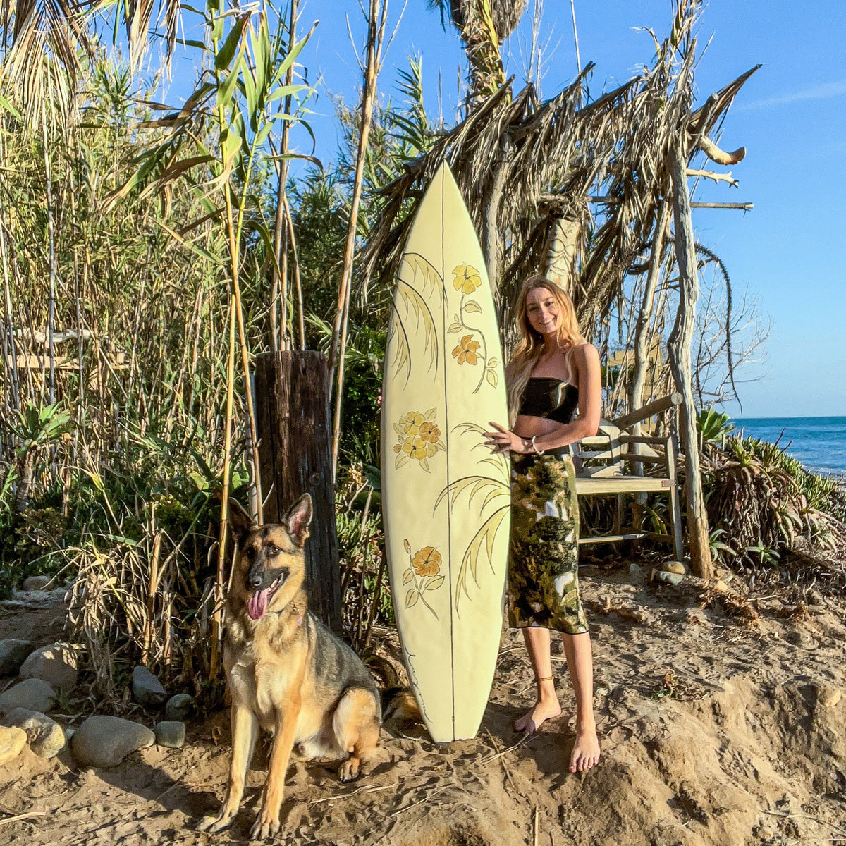 A woman stands on a sandy beach next to a cream-colored surfboard decorated with yellow floral designs, with a German Shepherd dog beside her. Palm trees, a wooden structure, and the blue ocean are visible in the background.