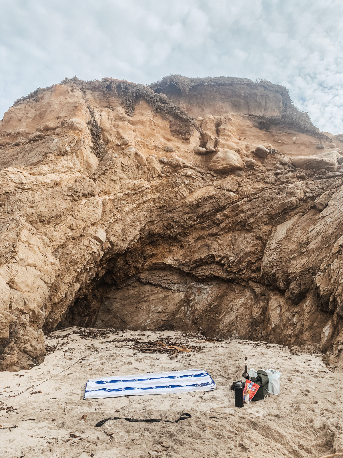 A sandy beach scene with a large tan rock cliff and cave formation behind it, under a cloudy sky. A blue-and-white striped towel, a black bag, and a small red-and-white item are placed on the sand near the base of the cliff.