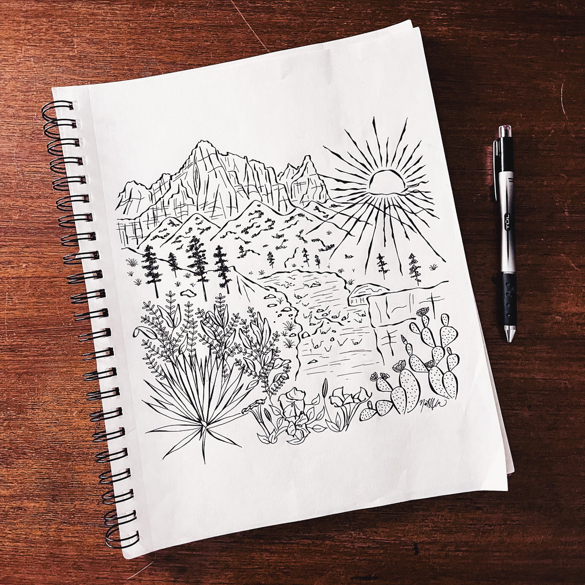 A spiral-bound sketchbook lying on a dark wooden table, featuring a black ink drawing of a mountain landscape with a sun, trees, and desert plants. A black and silver pen rests to the right of the notebook.