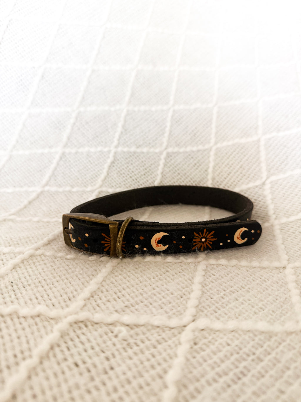 A black leather dog collar with celestial motifs including crescent moons and suns, displayed on a textured cream-colored fabric background.
