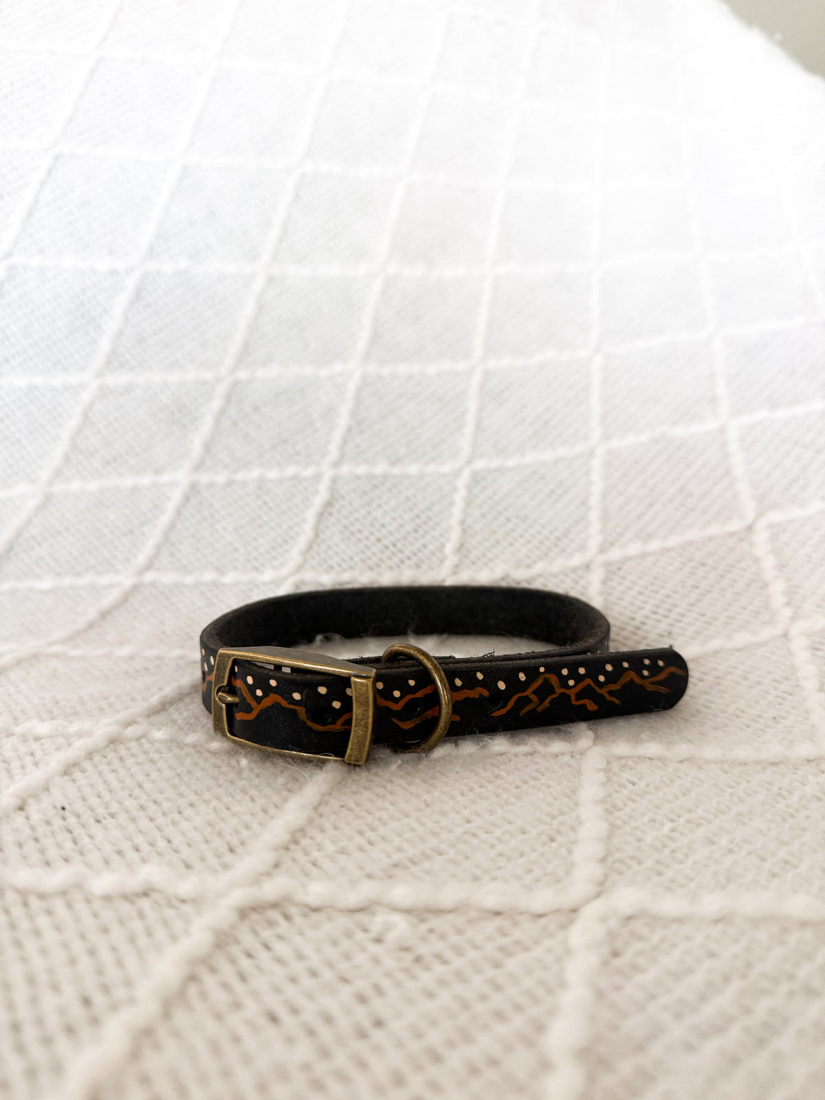 A black leather dog collar with hand-painted orange and white mountain patterns and a brass buckle, resting on a textured white fabric background.