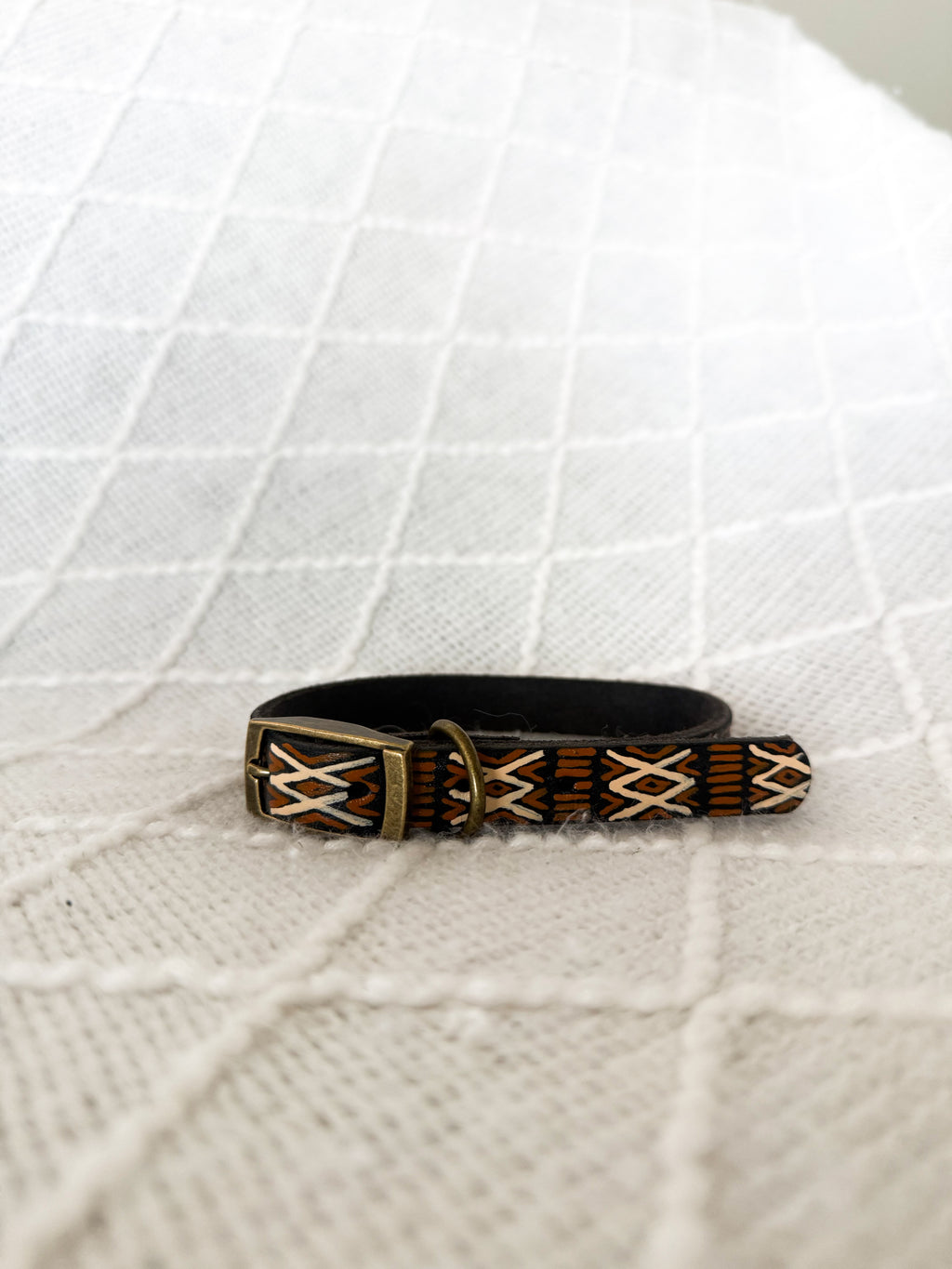 A black leather dog collar with brown and beige geometric patterned inlays and a brass buckle, resting on a white textured knit blanket.