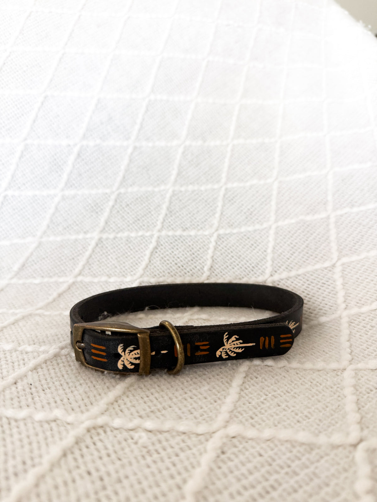 A black leather dog collar with gold-toned metal hardware and gold palm tree and stripe motifs, resting on a white textured knit blanket.