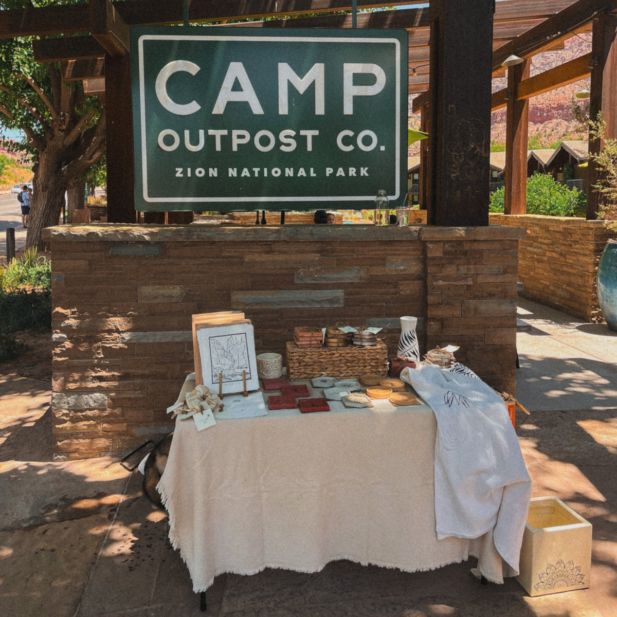 A rustic outdoor display table under a wooden structure with signs reading 'RESTAURANT OPEN DAILY' and 'CAMP OUTPOST CO. ZION NATIONAL PARK.