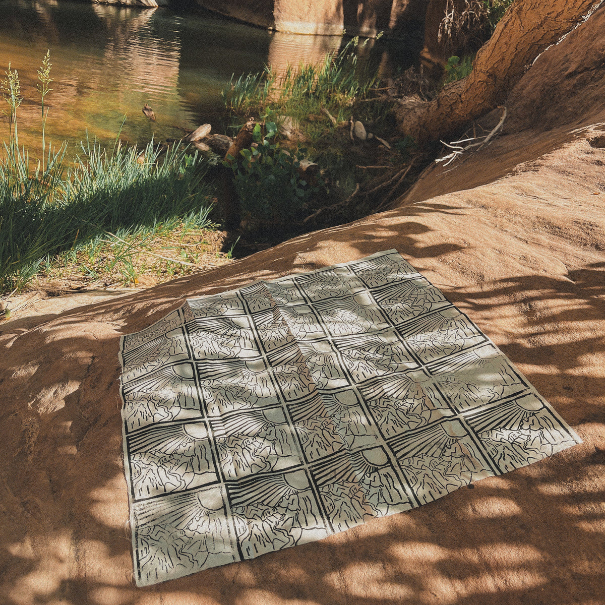 A rectangular outdoor rug with a geometric black and gray sunburst pattern is laid out on a sandy rock ledge next to a river canyon.