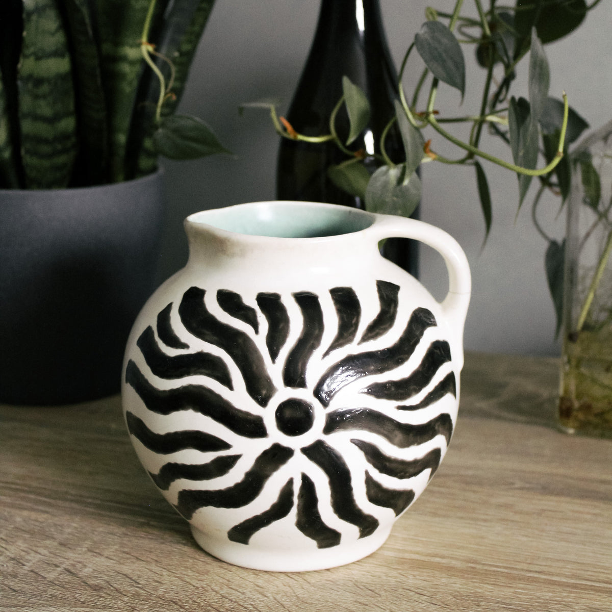 A white ceramic pitcher vase with a black hand-painted sunburst pattern, displayed on a wooden tabletop with potted plants in the background.