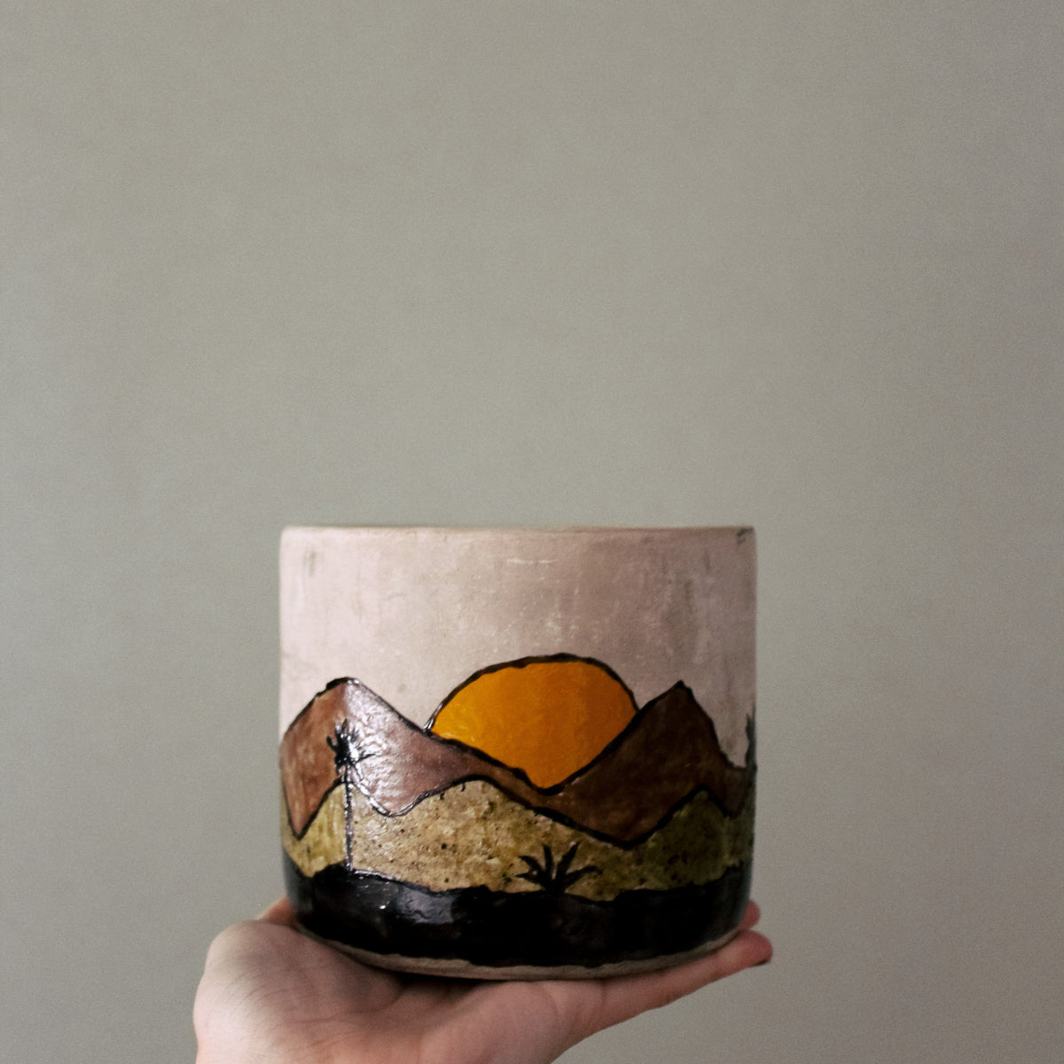 A hand holding a cylindrical ceramic planter with a hand-painted mountain and sun design in earthy tones against a plain gray background.