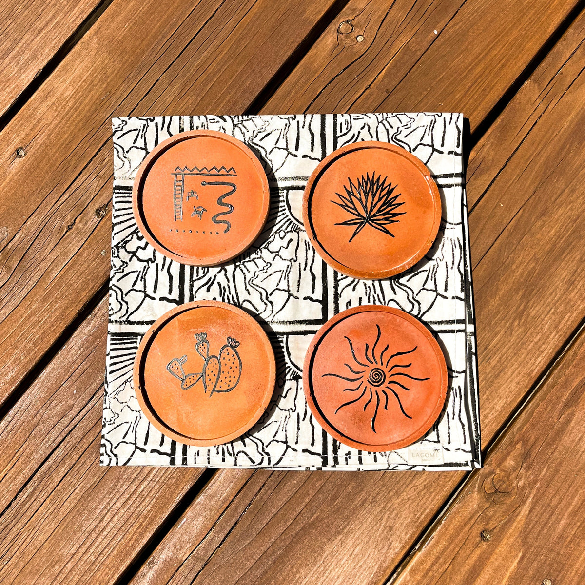 A square decorative board or tray with four circular orange coasters featuring black engraved symbols, placed on a wooden deck.