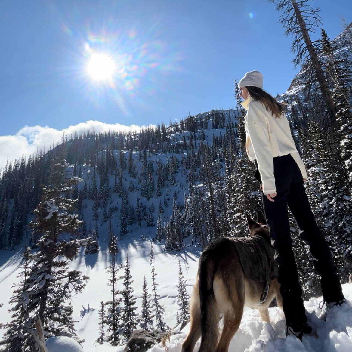 A woman in a white sweater and black pants stands in a snowy mountain landscape with evergreen trees under a bright blue sky with the sun shining. A dog is at her side.