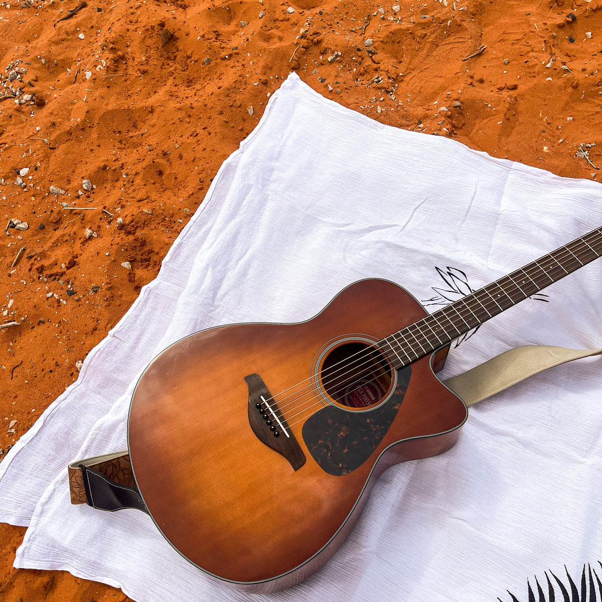 An acoustic-electric guitar with a natural sunburst finish and a black pickguard, resting on a white cloth over orange sand.