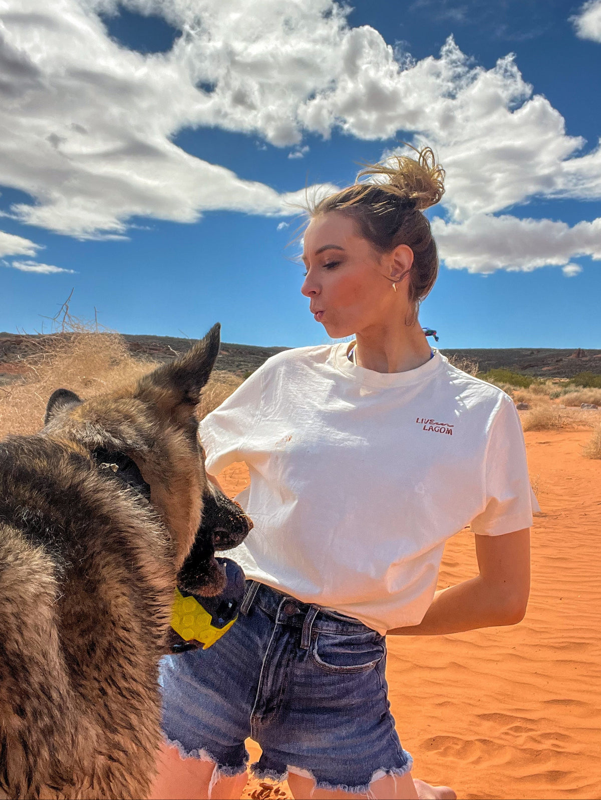 A woman in a white t-shirt and denim shorts stands in an orange desert landscape under a blue sky with clouds, interacting with a German Shepherd dog.