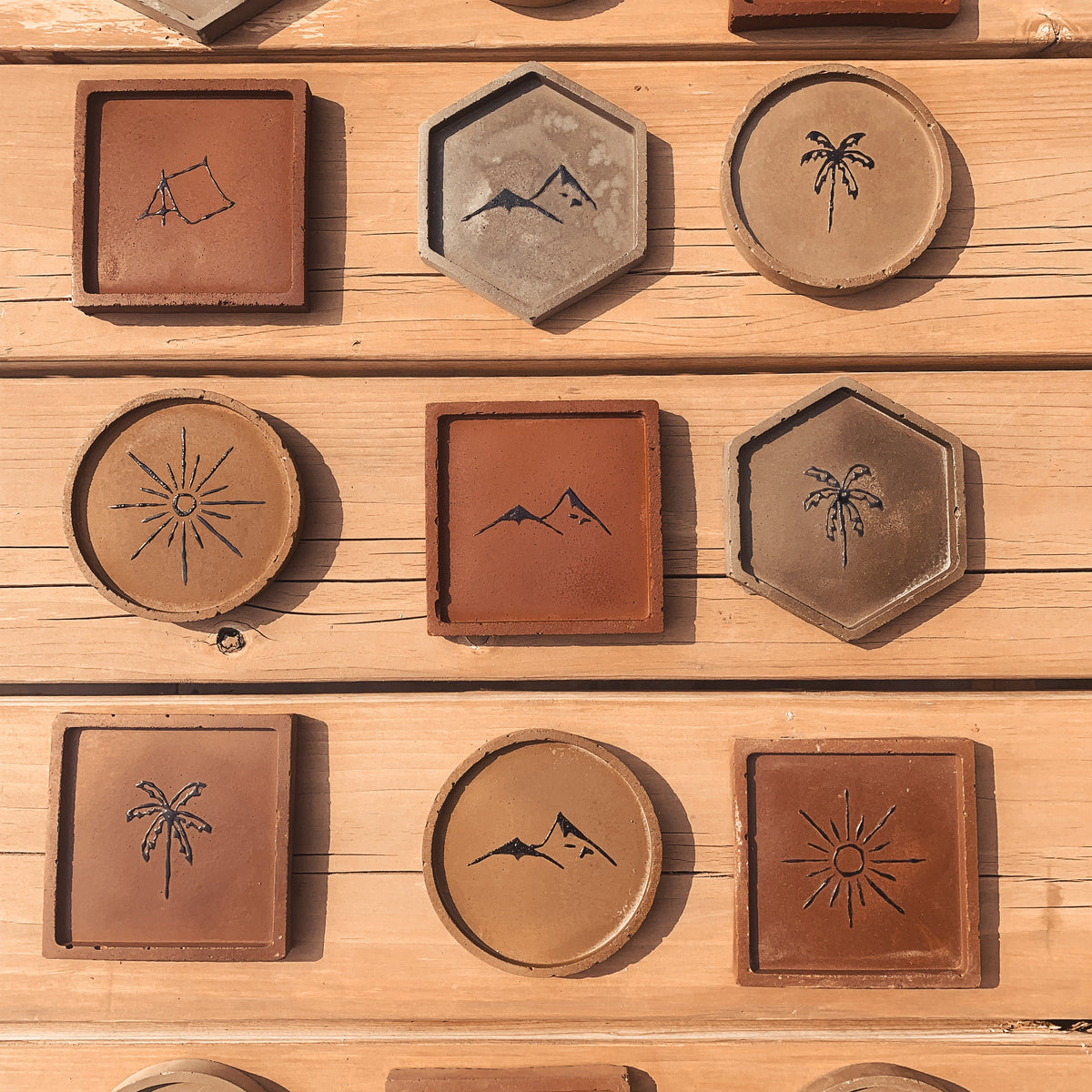 A collection of handmade terracotta coasters arranged neatly on a light wood plank background. The coasters come in square and hexagonal shapes, featuring various minimalist black engravings such as mountains, palm trees, suns, and waves.