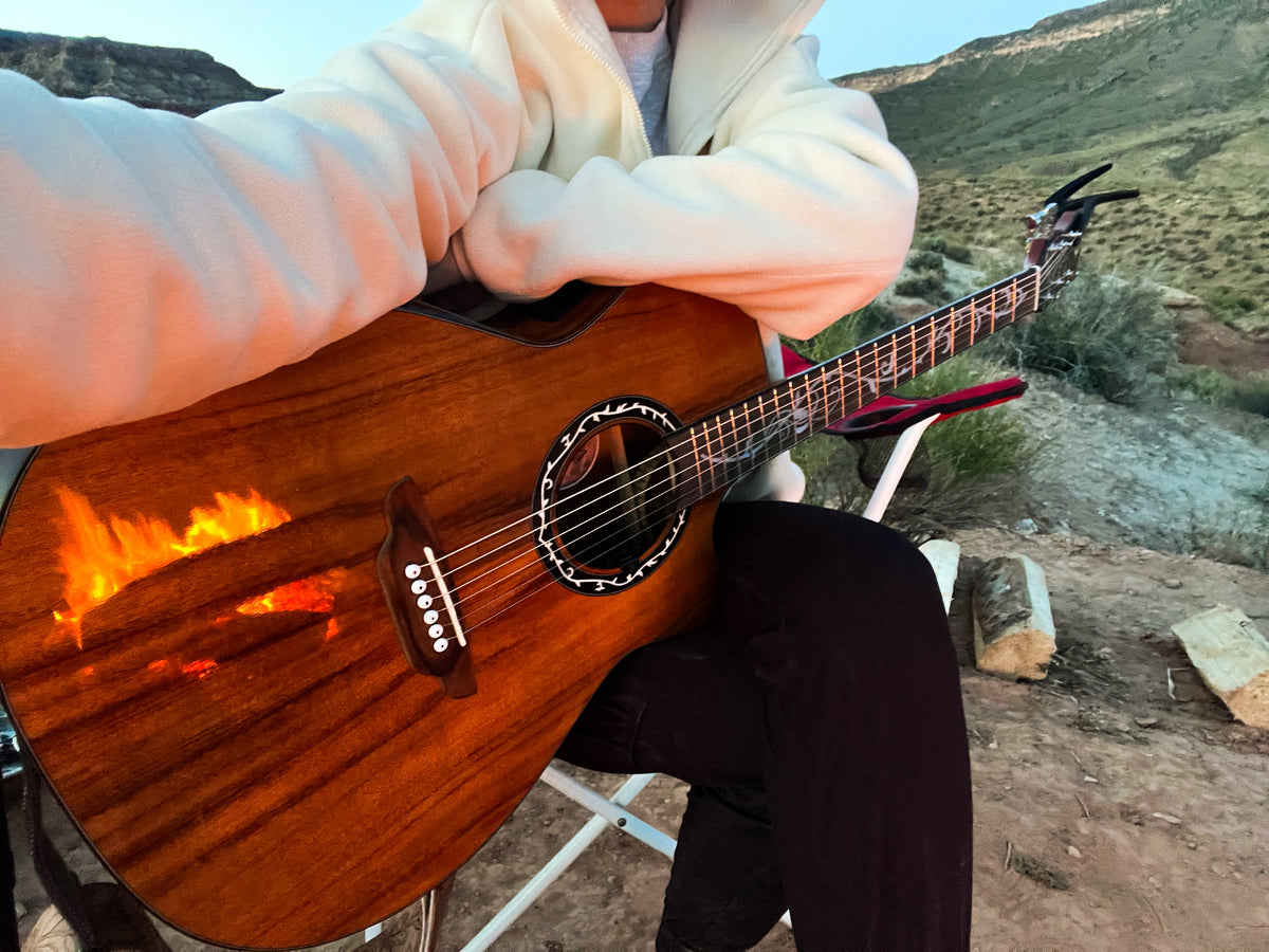 A person in a white hoodie and black pants sits outdoors holding a brown acoustic guitar with a flame-shaped graphic overlay on the lower left of the body, with a desert landscape in the background.