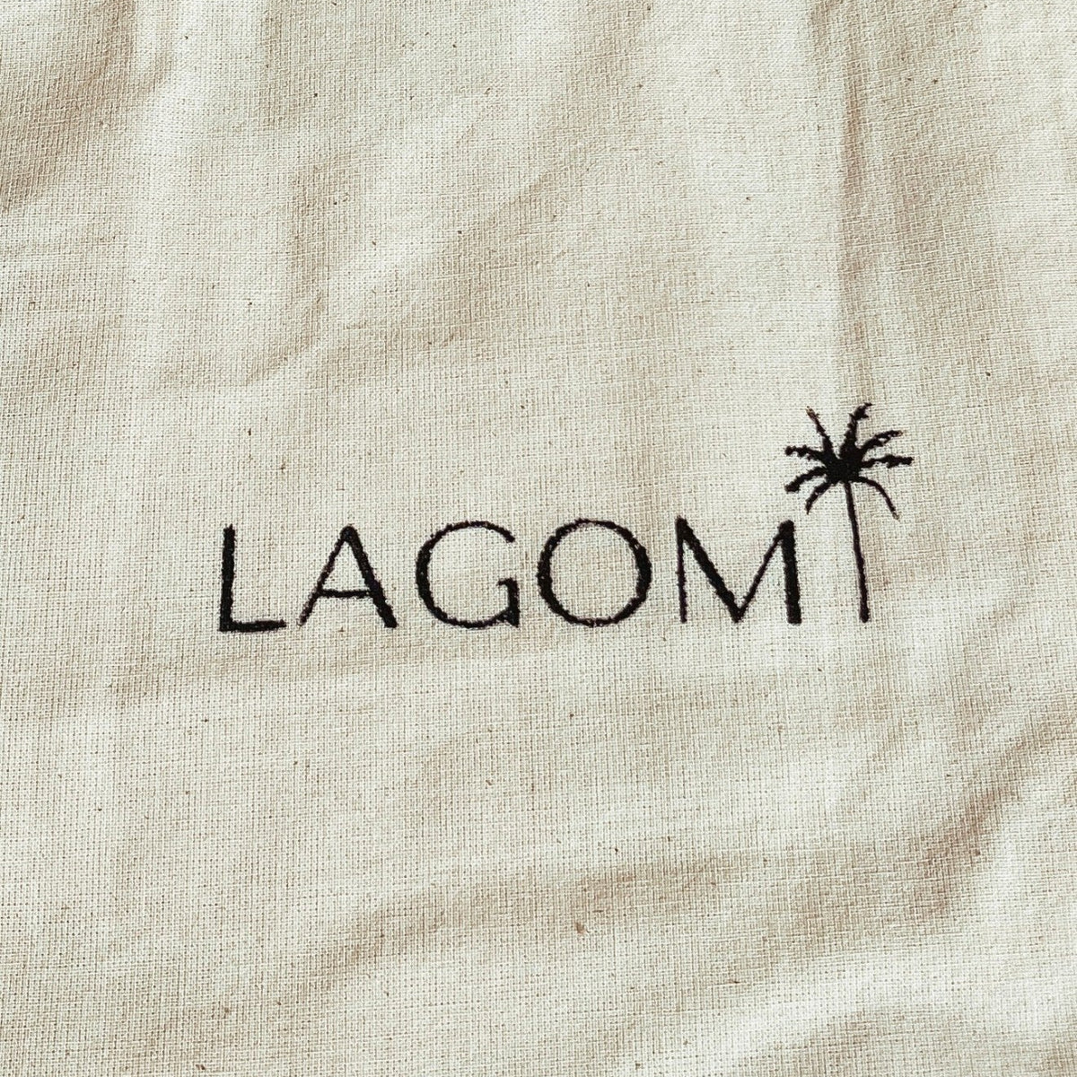 Close-up of a beige canvas or linen fabric with the word 'LAGOM' printed in black serif letters and a small black palm tree icon, showing the fabric texture and the printed logo clearly.